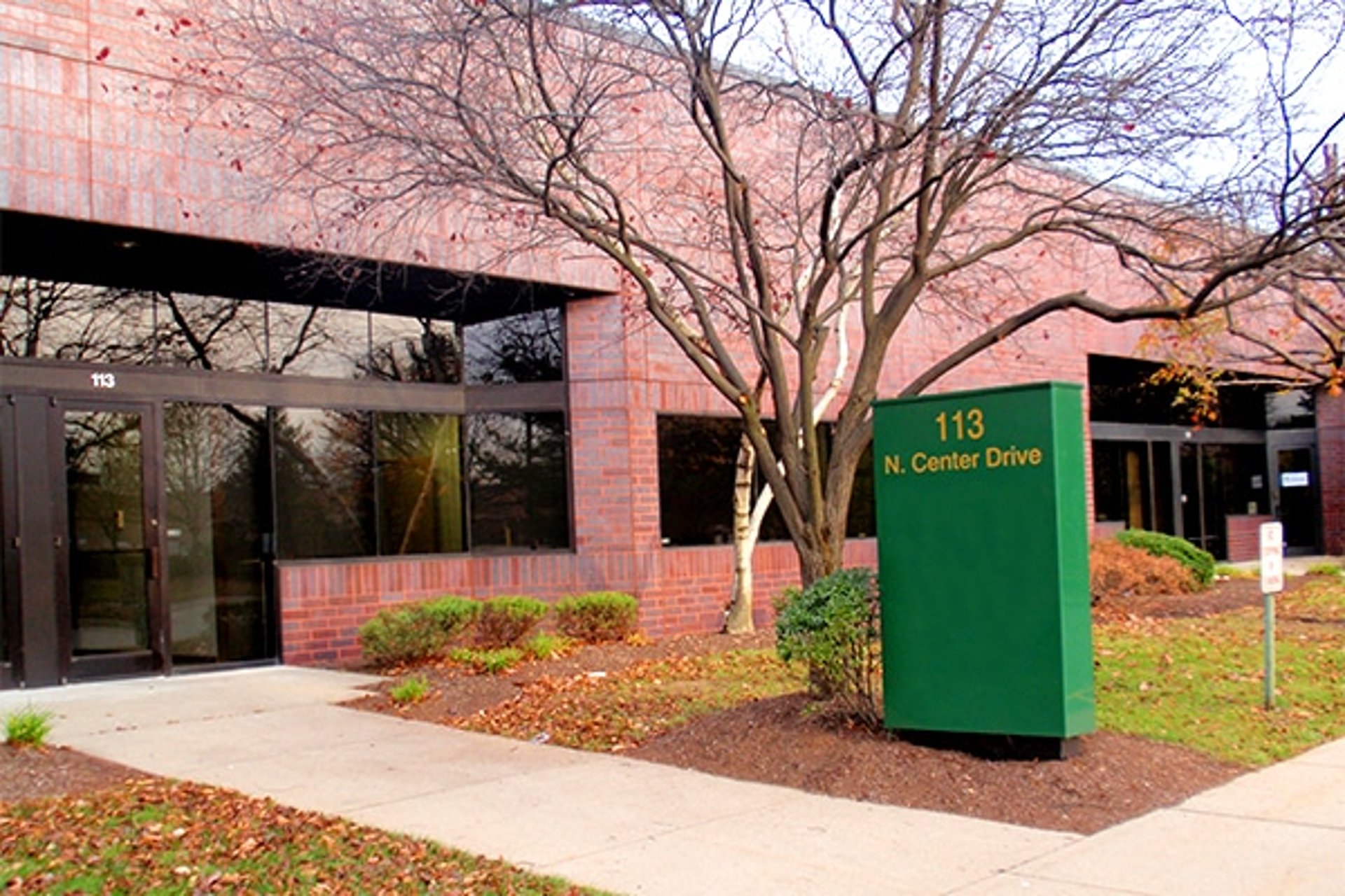 The front glass entrance of a long brick building with a winter tree with no leaves and a large green sign outside
