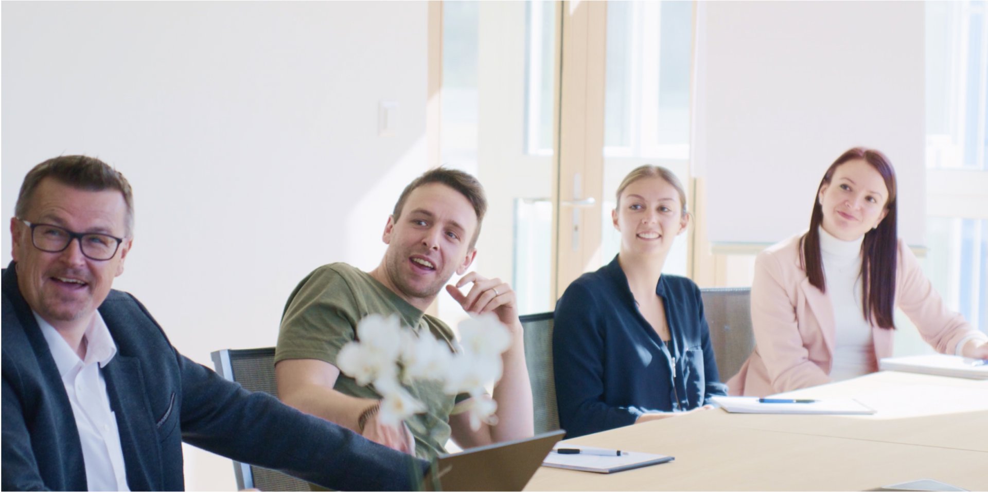 two men and two women sitting at a large wooden table with pens and paper in front of them in a white office room all looking at the same thing outside the shot