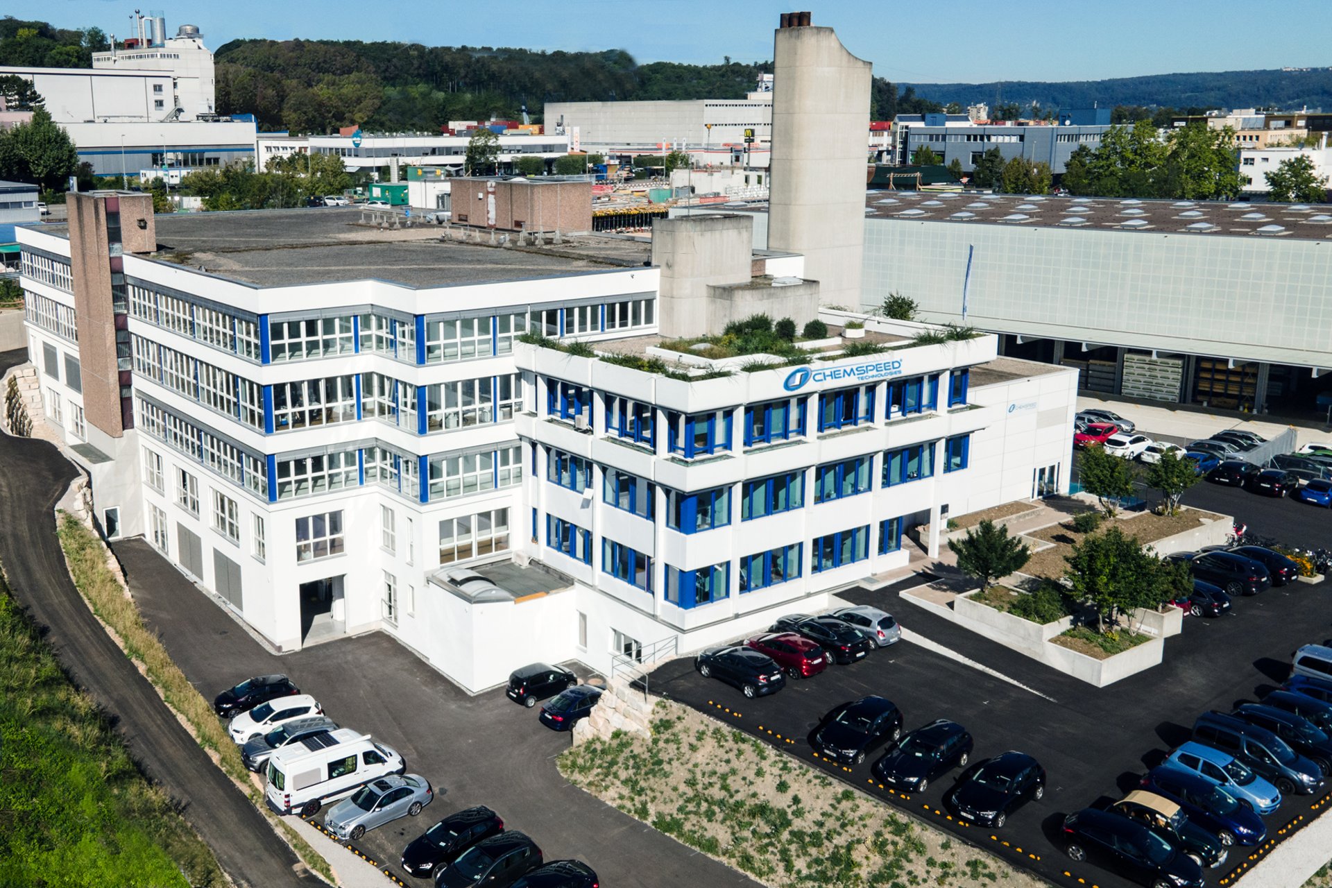 A drone shot of large white and blue building with cars parked around it in the car park and a blue sky