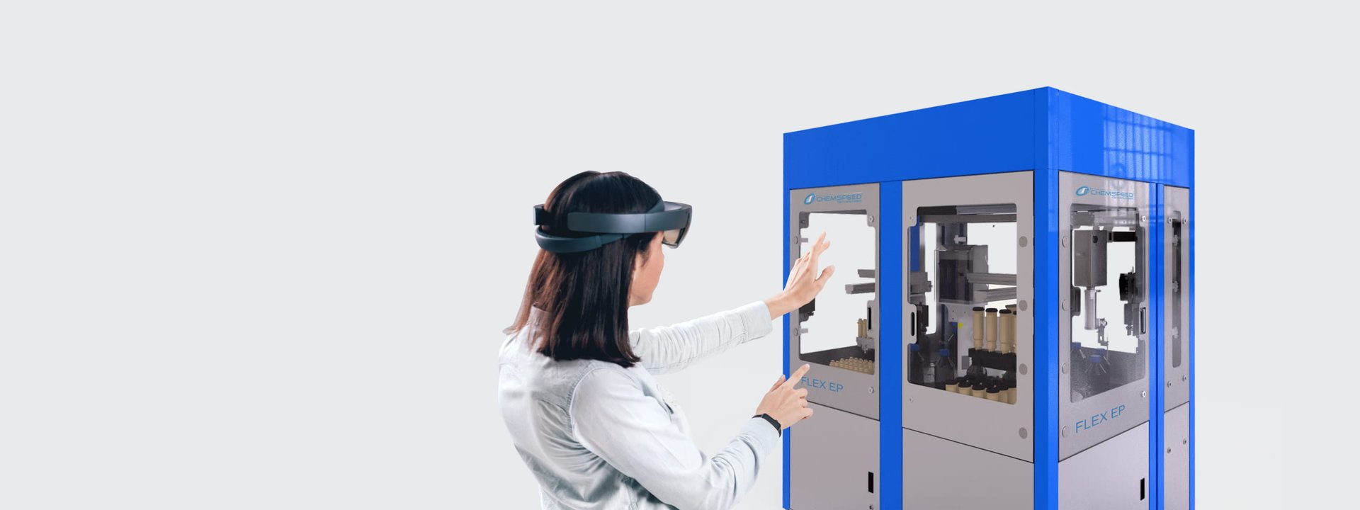 A female scientist with brown hair wearing an AR hololens and gesturing at a blue and grey Chemspeed robotic system