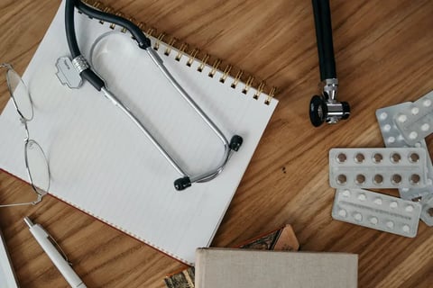 Medical professional's workspace with a stethoscope, eyeglasses, a pen, and a notepad on a wooden desk, alongside blister packs of pills and a closed book.