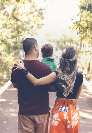 A family of three from behind, with a child on the father's shoulders, all holding hands, walking down a sunlit path surrounded by lush greenery.