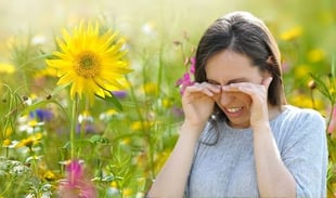 A woman rubbing her eyes in discomfort while standing in a field of blooming flowers, illustrating the symptoms of hay fever and seasonal allergies.