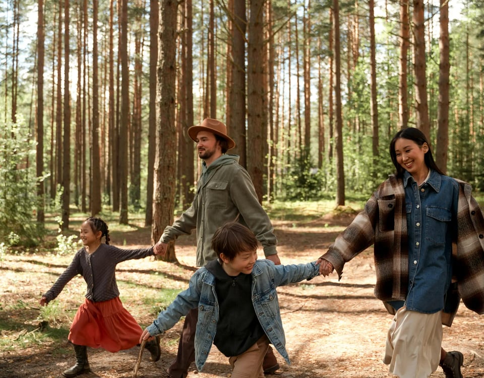 Family of four holding hands and walking through a pine forest, with two children playing and a man and a woman smiling joyfully.