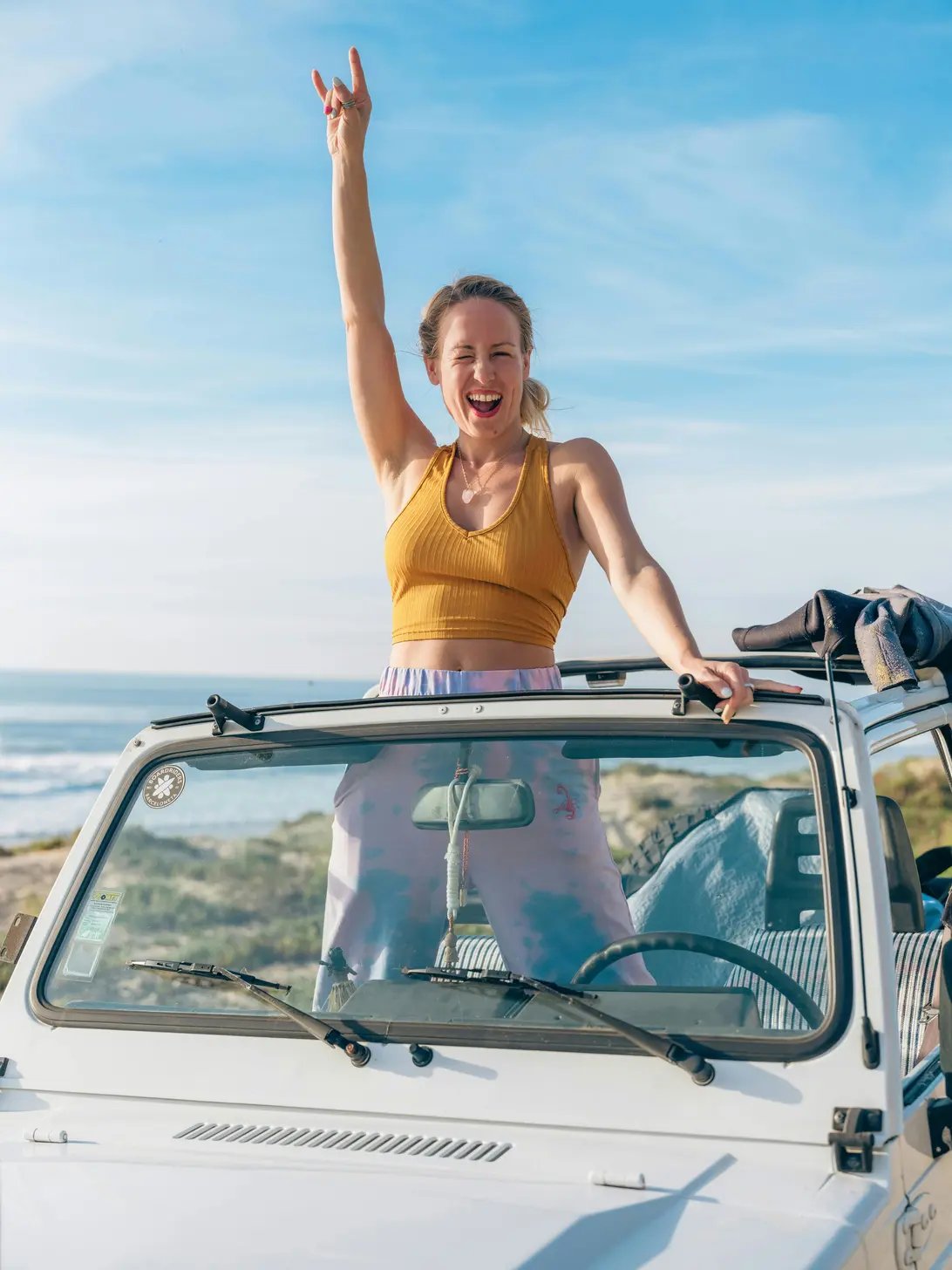Joyful woman in a yellow tank top standing through the sunroof of a white car, raising her hand in a peace sign, with a beach in the background.