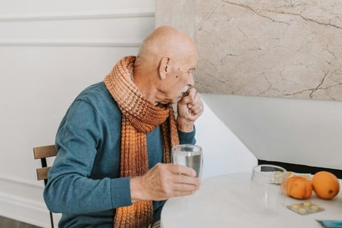 Senior gentleman coughing while sitting at a table, holding a glass of water.