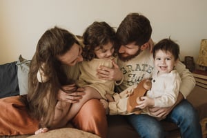 A young family consisting of a father, mother and two small children, cuddling on a couch in a mid-shot.