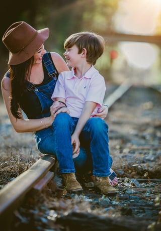 A woman in a brown hat sitting on a railway line holding her small son as they smile at each other.