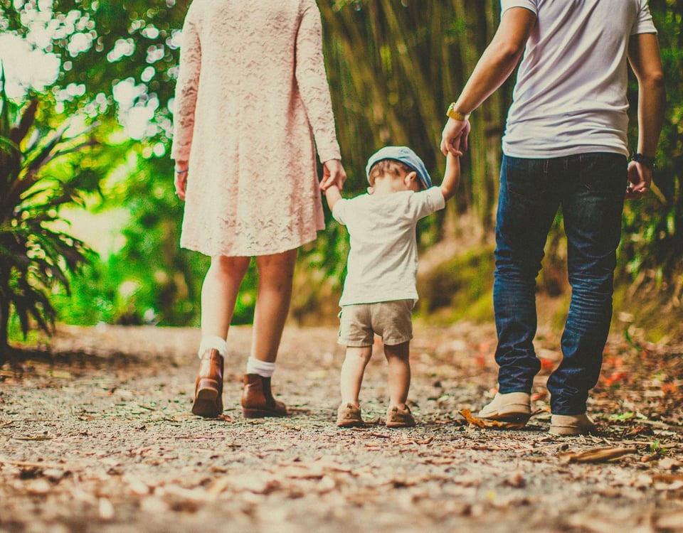 Mother and father walking holding hands with a toddler between them on a bushy path