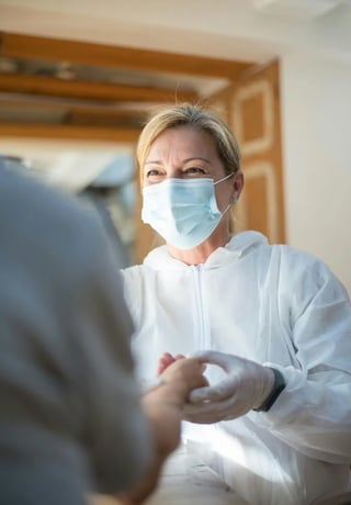 Compassionate female doctor in a protective mask and gown holding a patient's hand, offering comfort and reassurance in a clinic setting.