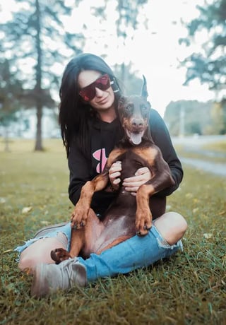 A woman in sunglasses sits on the grass cuddling her happy brown dog, with trees softly blurred in the background.