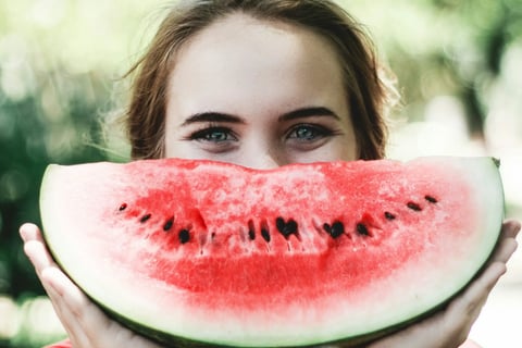 A young woman holding a slice of watermelon in front of her face like a smile.