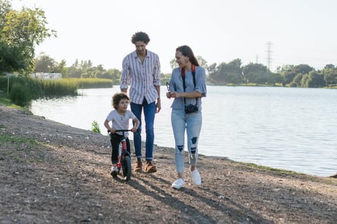 A man and woman walking a river bank with their small boy who is riding a scooter.