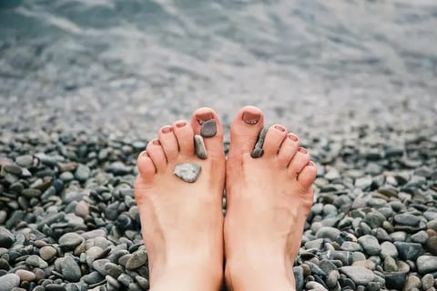 Bare feet with stones playfully placed on the toes, resting on a pebbly beach shore, with the water's edge in the background podiatry service.