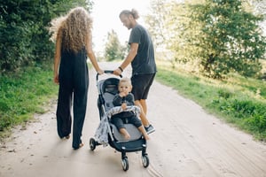A male and a female parents, with a toddler in a pram, walking on a country road.