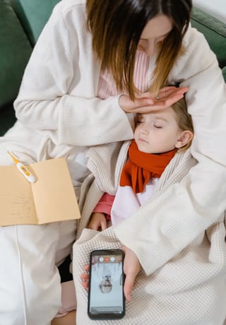 A concerned mother checking her young daughter's temperature, who is resting on a couch, with a thermometer and notepad nearby.