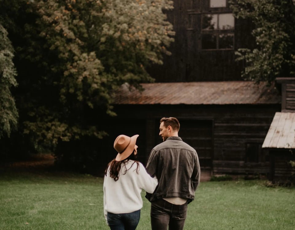 A couple walking hand in hand towards a rustic cabin surrounded by lush trees, with the woman wearing a wide-brimmed hat and the man in a casual jacket.