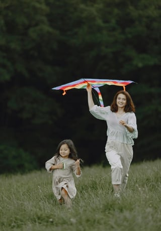 A young girl runs joyfully in a grassy field with a colorful kite flying behind her, guided by a smiling woman in a white outfit.
