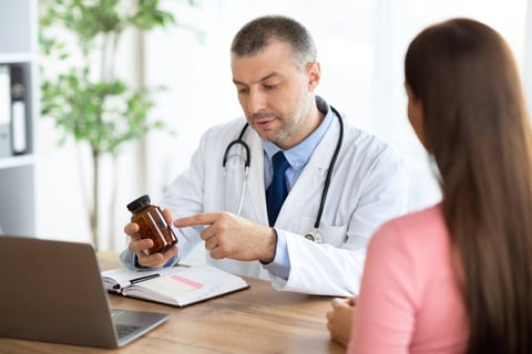 A male doctor holding a small brown bottle up to a women with brown hair who has her back turned to us.