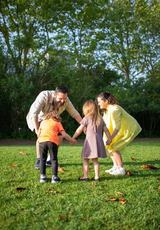Two parents and their children holding hands in a circle on a grassy lawn with fallen leaves, playing together in a sunny park with trees in the background.