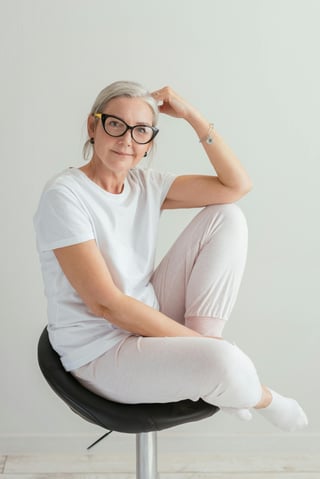 A poised woman with glasses sitting comfortably on a chair, dressed in white, with a thoughtful expression.