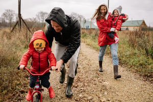 Two parents with two children, dressed in red, walking in the countryside. The father is pushing one toddler on a bike and the mother is carrying the other.