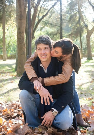 A smiling man seated on the ground covered with autumn leaves, with a woman hugging him from behind, both enjoying a sunny day in a park filled with trees
