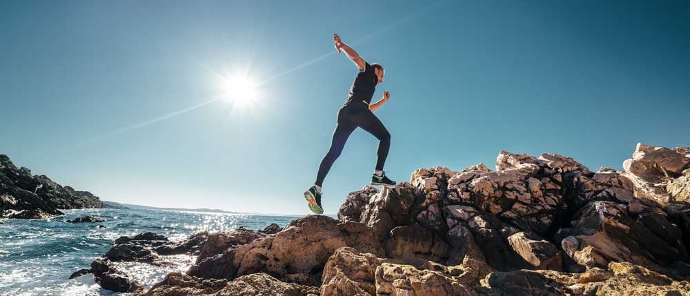 A person leaps across coastal rocks under a bright, clear sky, capturing a sense of adventure and a commitment to a healthy lifestyle.