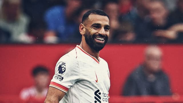 Mo Salah wearing a white Liverpool jersey and smiling in joy/disbelief