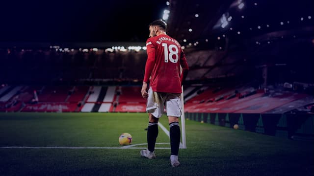 Bruno Fernandes taking a corner. In the image, he's wearing a red Manchester United flag and standing by the corner flag with the stadium stretching off into the background