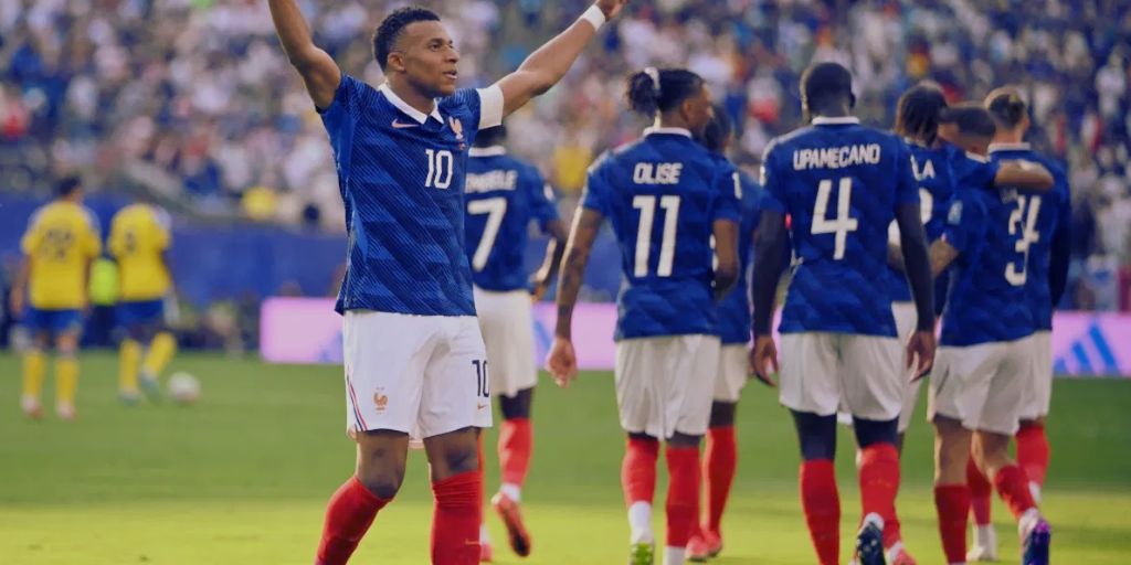 Kylian Mbappe celebrating a goal while his teammates walk away from the camera. Two blurrry Sweden players also shown in the background