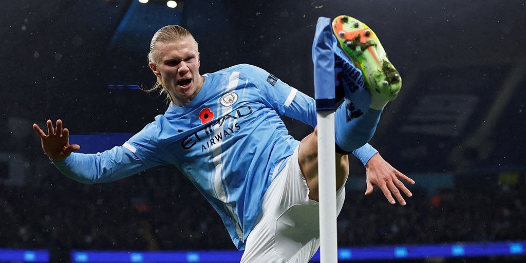Erling Haaland celebrating a goal by kicking the white and blue corner flag while dressed in a light blue Manchester City top, white shorts, light blue socks and lime green boots