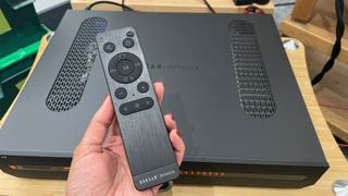 Telecomanda Roksan Atessa Streaming Amplifier ținută în mână deasupra unității amplificatorului