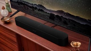 Un Sonos Beam Gen 2 negru în fața unui televizor pe un suport de lemn