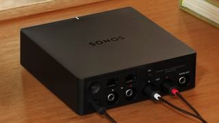 Un Sonos Port negru pe o masă de lemn, cu trei cabluri conectate la el.