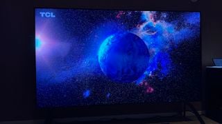 Televizor Mini LED TCL 115QM89 fotografiat într-o cameră de hotel. Pe ecran este imaginea Pământului și o constelație strălucitoare.