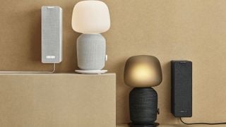 Două difuzoare lampă de masă Sonos IKEA Symfonisk (una albă, una neagră) una lângă alta pe o suprafață bej în fața unui perete bej. Difuzorul alb este ridicat mai sus decât cel negru.