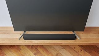 Bara de sunet Sony HT-SF150 poziționată pe un cabinet de lemn, între picioarele unui TV
