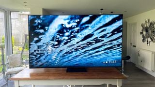 Televizorul TCL 85C805K 4K pe o masă într-o cameră cu o grădină vizibilă printr-o fereastră în fundal. Pe ecran este un prim-plan al unei suprafețe de apă.