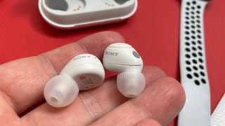 Căști wireless Sony WF-C700N albe în mână peste o masă roșie, cu cutia lor parțial vizibilă în fundal.