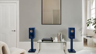 O pereche de KEF R3 Meta montate pe suport în ediție specială indigo gloss, flancând o masă de cafea din sticlă într-o cameră albă.