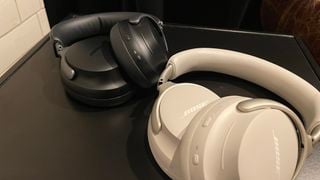 Două perechi de căști Bose QuietComfort Ultra (una neagră, una albă) pe o suprafață neagră lângă un perete cu faianță albă.