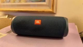 O boxă JBL Flip 5 albastră pe o suprafață mov în fața unui tablou agățat de perete.