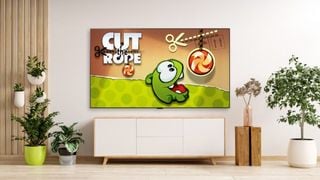 Ecranul de start al jocului Cut the Rope este pe televizor într-o cameră luminoasă. Sunt plante de apartament pe podea și sertare sub televizor.