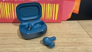 Căști in-ear JBL Live Beam 3, una în carcasă și cealaltă lângă, pe o poliță din lemn