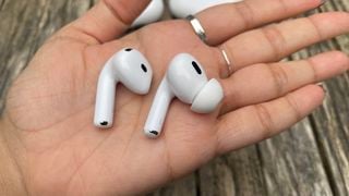 O cască din fiecare model Apple AirPods Pro 2 și AirPods 4 în palma cuiva în fața unei suprafețe de lemn.