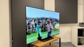 Televizor LG C4 de 65 de inci fotografiat dintr-un unghi ușor pe un suport de lemn. Pe ecran este afișată o imagine cu doi jucători de golf felicitându-se reciproc.
