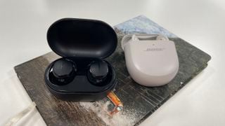 Bose QuietComfort Earbuds Ultra și QuietComfort Earbuds (2024) în husele lor de încărcare pe o placemat gri.
