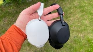 O mână ținând căștile albe Sonos Ace și negre Sony WH-1000XM5 over-ear de benzile de cap în fața unui petic de iarbă.