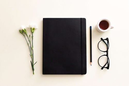 a black notebook with glasses pencil and a pencil next to a cup of tea